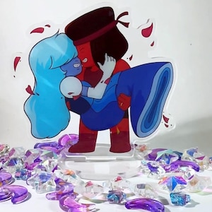 Puede incluir: Un soporte acrílico transparente con una ilustración de dibujos animados de dos personajes abrazándose. Uno es rojo, el otro azul. El soporte está rodeado de decoraciones cristalinas transparentes y moradas. Pétalos rojos dispersos.