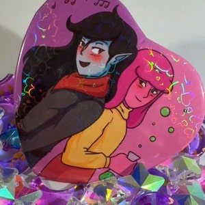 May include: A pink heart-shaped button with a holographic finish features a cartoon couple. The woman has pink hair and is wearing a yellow sweater. The man has black hair and is wearing a red scarf. The background is pink with holographic hearts and music notes.