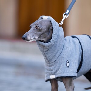 May include: A grey dog coat with a high collar and a black leash attachment point. The garment features a textured heather grey fabric, a small circular logo patch on the side, and black trim along the edges. The coat covers the torso and neck.