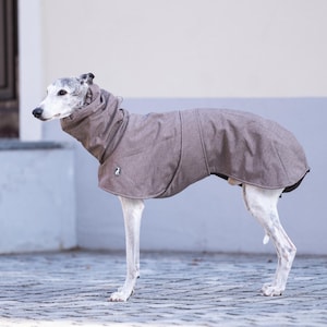Pu&ograve; includere: Un cappotto per cani color grigio-marrone con collo alto e una piccola toppa circolare con logo sul lato. Il capo presenta un design strutturato che copre la schiena e il petto, realizzato per una corporatura canina snella e liscia.