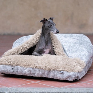 May include: A grey Italian Greyhound dog resting inside a rectangular pet bed. The bed features a grey textured exterior fabric and a soft, beige faux-fur interior lining that forms a hood-like cover over the dog. Two small logo tags are visible.