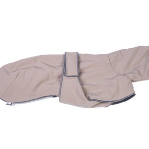 May include: A beige dog coat featuring a smooth, water-resistant outer shell with dark gray trim along the edges. The garment includes a wide, adjustable strap across the midsection with a hook-and-loop closure and a small white brand tag.