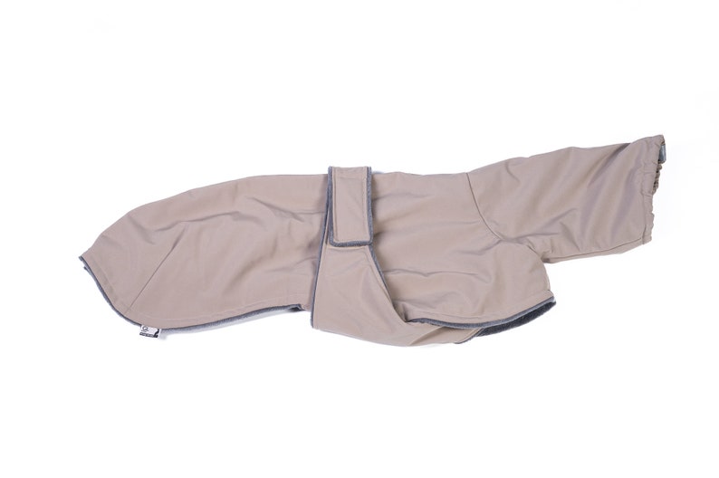 May include: A beige dog coat featuring a smooth, water-resistant outer shell with dark gray trim along the edges. The garment includes a wide, adjustable strap across the midsection with a hook-and-loop closure and a small white brand tag.
