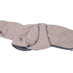 May include: A beige dog coat featuring a smooth outer shell with a dark gray trim and a soft, dark gray interior lining. The garment includes a wrap-around belly strap with a small circular logo emblem on the side and an elasticized neck opening.