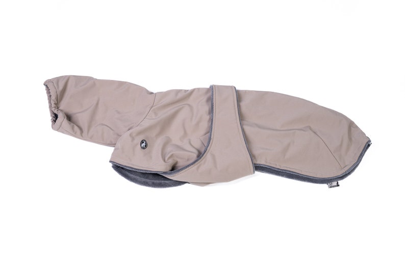 May include: A beige dog coat featuring a smooth outer shell with a dark gray trim and a soft, dark gray interior lining. The garment includes a wrap-around belly strap with a small circular logo emblem on the side and an elasticized neck opening.