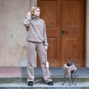 Może przedstawiać: Kobieta w dopasowanym zestawie w kolorze taupe: golf i spodnie z warkoczowym wzorem. Mały pies w pasującym swetrze na smyczy. Tło obejmuje drewniane drzwi i kamienną fasadę.
