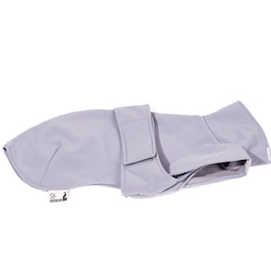 May include: A light gray pet coat featuring a smooth exterior, a structured collar area, and a wide adjustable strap across the midsection with a hook-and-loop closure. A small white rectangular tag with black text is attached to the lower edge.