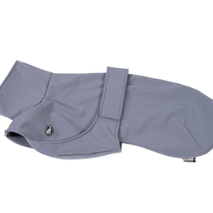 May include: A solid light gray dog coat featuring a smooth, matte finish, a high collar, and a vertical strap closure across the midsection. A small, circular black and white logo patch depicting a bird is visible on the side of the garment.