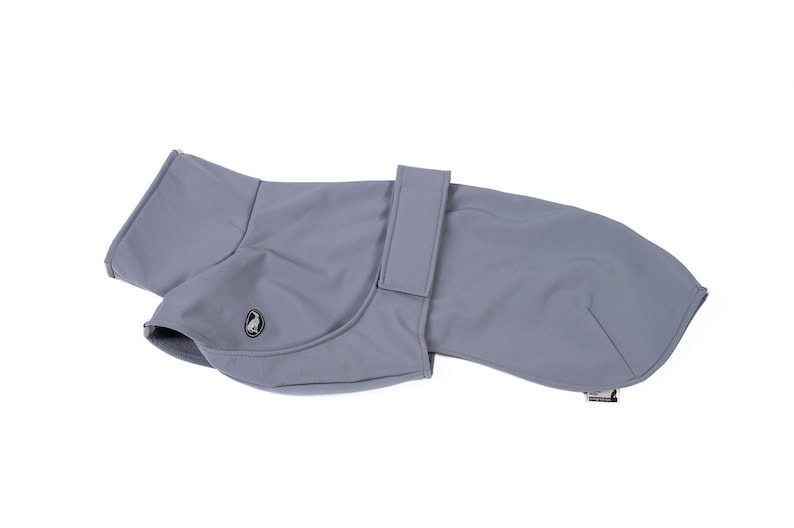 May include: A solid light gray dog coat featuring a smooth, matte finish, a high collar, and a vertical strap closure across the midsection. A small, circular black and white logo patch depicting a bird is visible on the side of the garment.