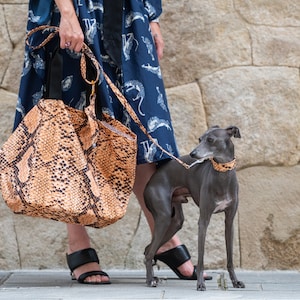 May include: A large tote bag with a snakeskin pattern in shades of brown and black is held by a person. A grey Italian Greyhound dog wears a matching collar and leash. The dog stands on a paved surface next to a stone wall.