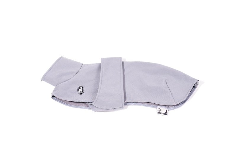 May include: A light gray dog coat featuring a smooth, solid-colored exterior, a high collar, and a wide adjustable strap across the midsection. A small circular logo with a dog silhouette is on the side, and a rectangular brand tag is attached.