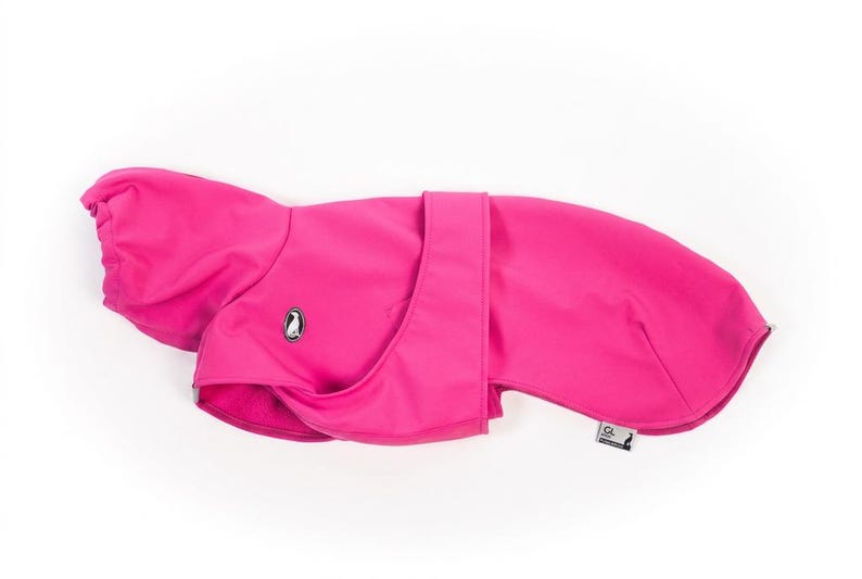 Pu&ograve; includere: Un cappotto per cani rosa acceso caratterizzato da un esterno liscio in tinta unita, una zona del colletto strutturata e una chiusura centrale a cinturino. Sul lato &egrave; visibile un piccolo logo ovale con un pinguino, insieme a una piccola etichetta.