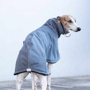 May include: A grey dog coat with a high collar and black trim, featuring a textured fabric finish and a small rectangular tag at the hem. The garment covers the back and neck of a brindle and white dog wearing a metal collar with a ring attachment.
