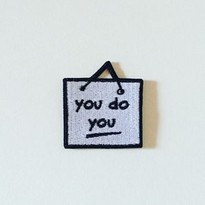 May include: A black and white embroidered patch with the text "you do you" on a white background. The patch has a black hanging loop at the top.