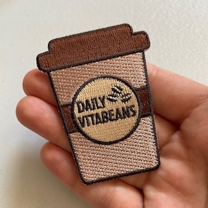 Könnte beinhalten: Ein gestickter Aufnäher in Braun und Beige, der eine Kaffeetasse mit dem Text "Daily Vitabeans" und einer Kaffebohnengrafik zeigt.