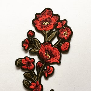 Op de afbeelding: Rode geborduurde bloemenpatch met groene bladeren en stelen. De bloemen zijn in verschillende maten en vormen, sommige hebben een meer open bloei dan andere.