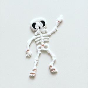 May include: White embroidered skeleton patch with black eyes and pink accents. The skeleton is waving with one arm raised.