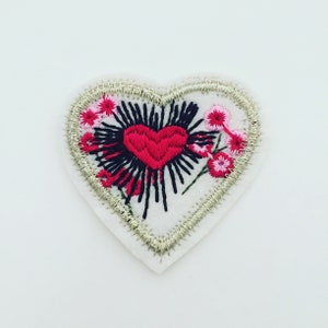 Könnte beinhalten: Ein weißer Filz-Herz-Patch mit einem roten gestickten Herzen in der Mitte. Das Herz ist umgeben von schwarzen gestickten Linien und rosa und weißen gestickten Blumen.