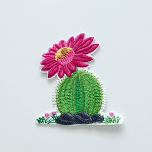 Pode incluir: Etiqueta bordada para passar a ferro com uma flor de cacto rosa com um cacto verde e pedras pretas.