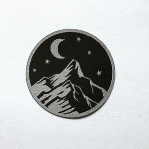 Könnte beinhalten: Ein gestickter Patch in Schwarz und Silber mit einer Silhouette einer Bergkette unter einem Halbmond und Sternen.