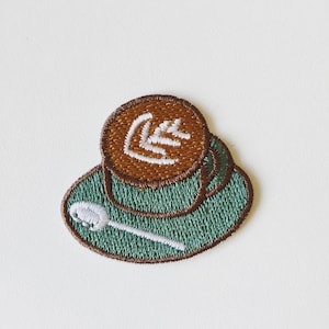 Może przedstawiać: Naszywka haftowana przedstawiająca filiżankę kawy z łyżeczką i wzorem latte art w odcieniach zieleni i brązu.