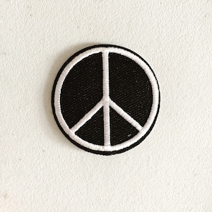 Może przedstawiać: Czarna naszywka z symbolem pokoju z białym obramowaniem. Naszywka jest okrągła i ma fakturowaną powierzchnię.