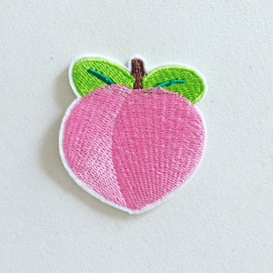 Könnte beinhalten: Ein gestickter Pfirsich in Rosa mit grünen Blättern. Der Pfirsich ist eine beliebte Frucht und ein Symbol des Sommers.