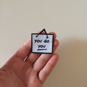 May include: White embroidered patch with black stitching. The patch is shaped like a square with rounded corners and has a black string loop at the top. The text on the patch reads "you do you".