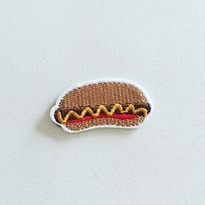 May include: A brown and white embroidered patch of a hot dog in a bun with yellow mustard and red ketchup.