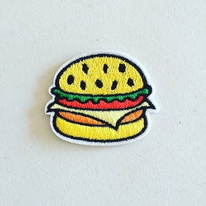 Pode incluir: Um remendo bordado amarelo de um hambúrguer de desenho animado com alface verde, tomate vermelho e queijo laranja.