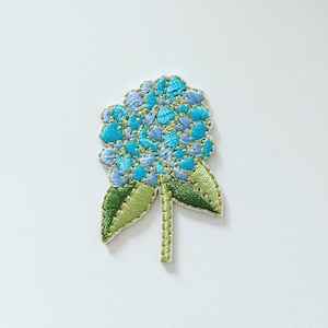 Op de afbeelding: Een geborduurde bloemenpatch in blauw en groen met een gouden rand. De bloem heeft een cluster van kleine blauwe bloemblaadjes en groene bladeren.