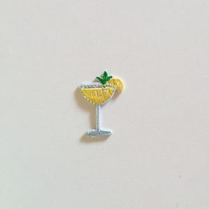 Op de afbeelding: Geborduurde patch van een gele cocktail in een glas met steel, gegarneerd met een groene tak en een schijfje citroen. De patch heeft een lichtblauwe steel en basis, tegen een witte achtergrond.