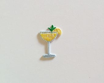 May include: Embroidered patch of a yellow cocktail in a stemmed glass, garnished with a green sprig and a lemon slice. The patch has a light blue stem and base, and is set against a white background.