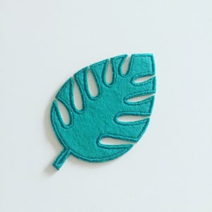May include: A teal green felt monstera leaf with a stitched outline. The leaf is a simple shape with a rounded tip and a single stem.