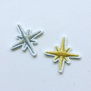 May include: Two embroidered star-shaped patches, one silver and one gold, on a white background.