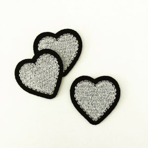 May include: Three silver heart-shaped patches with black outlines. The hearts are made of a textured fabric.