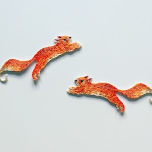 Può includere: Due scoiattoli arancioni ricamati con code bianche, uno salta in alto e l'altro salta in basso.