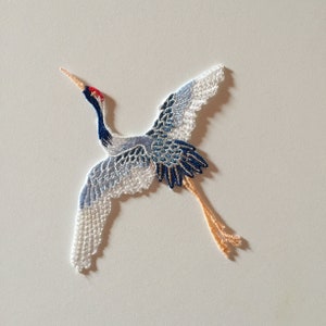 May include: A white, blue, and red embroidered crane with outstretched wings. The crane is made of delicate lace and has a long, thin neck.