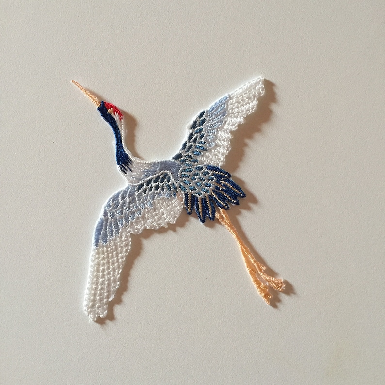 May include: A white, blue, and red embroidered crane with outstretched wings. The crane is made of delicate lace and has a long, thin neck.