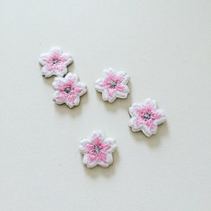 Könnte beinhalten: Fünf gestickte Blumenpatches in Rosa und Weiß. Die Blumen haben eine weiße Mitte mit einem rosa Umriss und einer silbernen Mitte.