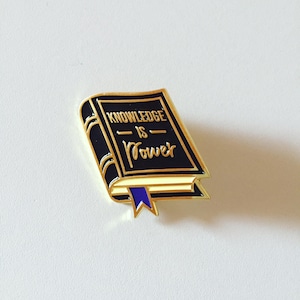 Könnte beinhalten: Goldfarbener, buchförmiger Emaille-Pin mit schwarzem Umschlag und einem blauen Band als Lesezeichen. Der Umschlag trägt die Aufschrift "Knowledge is Power".