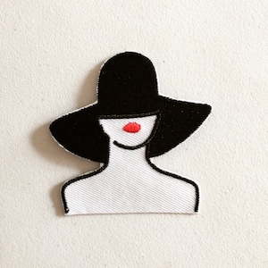 Può includere: Toppa ricamata raffigurante una donna stilizzata con un cappello a tesa larga. Il design utilizza nero, bianco e rosso. Il cappello e i capelli della donna sono neri, le sue labbra sono rosse e il contorno è nero.