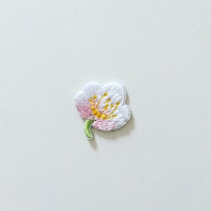 May include: An embroidered flower patch in white with pink and yellow accents. The flower has five petals and a green stem.