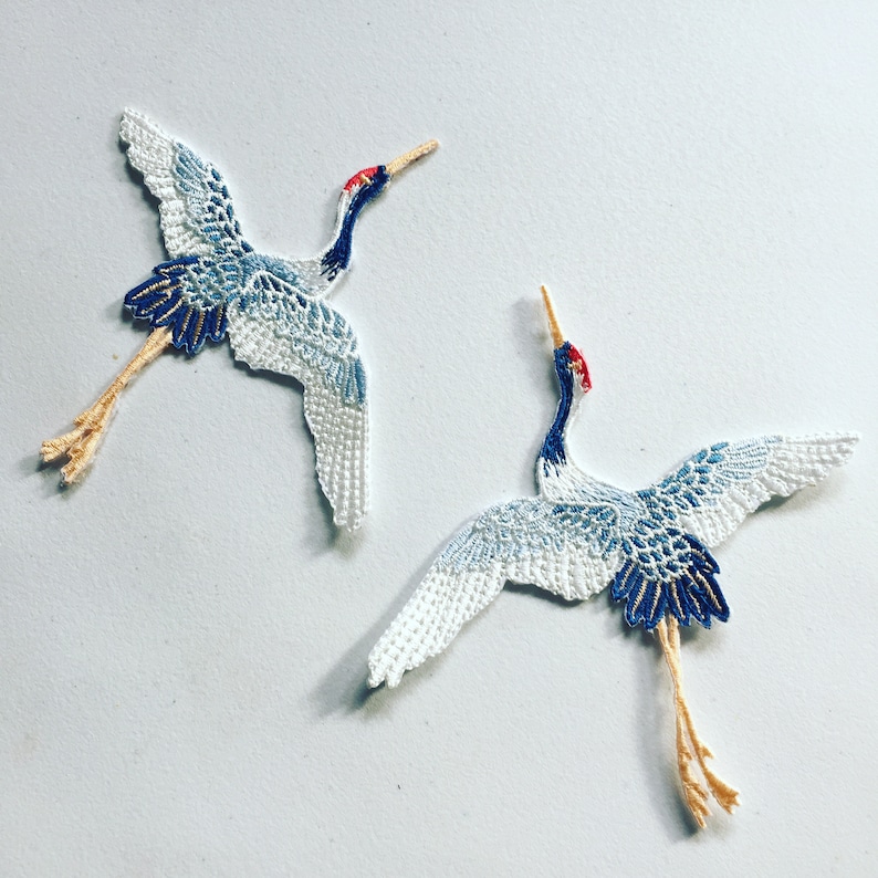 May include: Two white and blue embroidered crane appliques with red beaks and yellow legs. The cranes are in a flying position with their wings spread out.