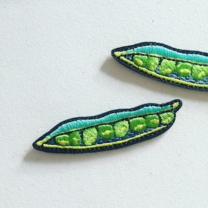 May include: Two embroidered iron-on patches shaped like pea pods. The pea pods are green with a light blue outline and yellow stitching. The peas inside are green with a yellow outline.
