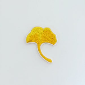 May include: A yellow embroidered ginkgo leaf patch. The leaf is a light yellow color and has a stem that curves to the right.