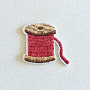 May include: Embroidered iron-on patch of a spool of red thread with a brown wooden spool and a brown thread tail.