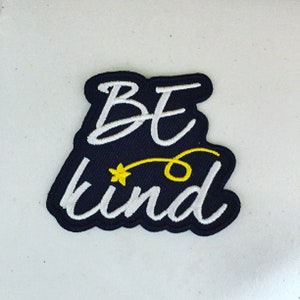 May include: Black iron-on patch with white embroidered text that reads "Be kind" with a yellow star and a yellow swirl.