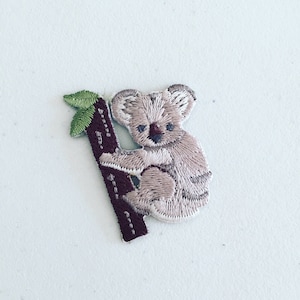 May include: Embroidered iron-on patch of a koala bear climbing a tree. The koala is brown and white, and the tree is brown with green leaves.
