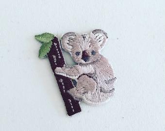 May include: Embroidered iron-on patch of a koala bear climbing a tree. The koala is brown and white, and the tree is brown with green leaves.